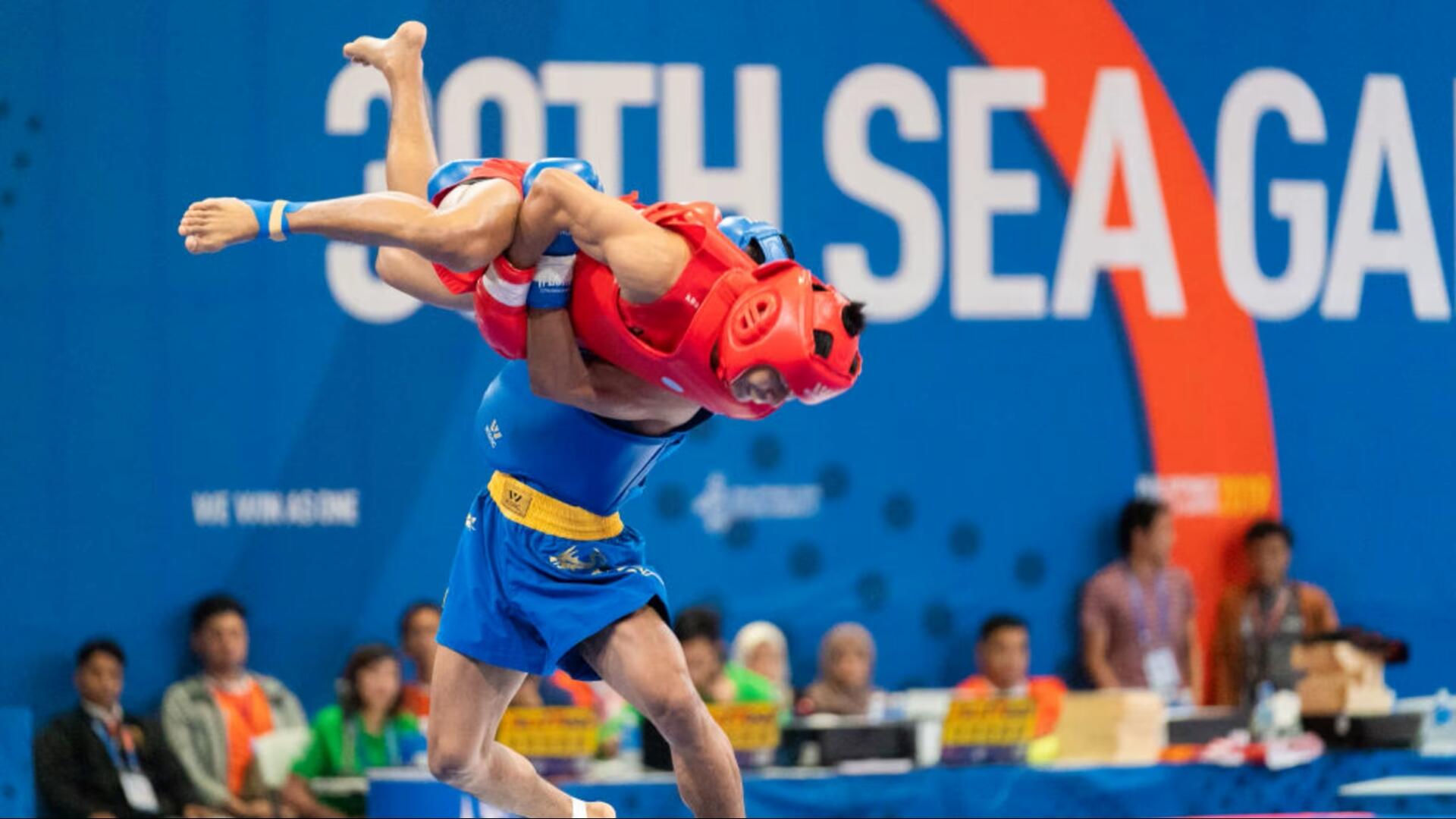 Two martial artists in protective gear compete in a wrestling move at the 30th SEA Games, with spectators blurred in the background.
