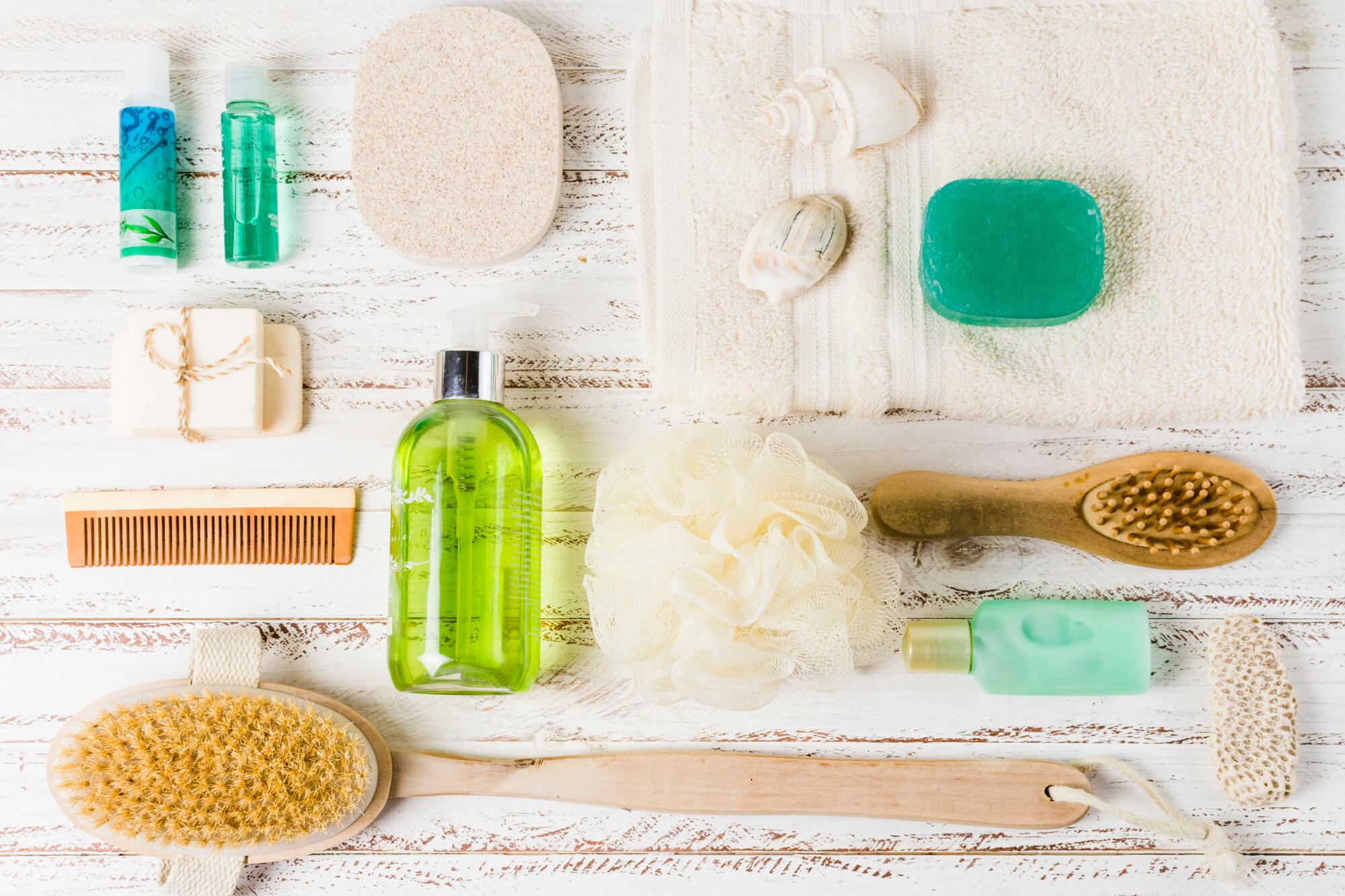 Assorted bath items including brushes, soap bars, lotions, a loofah, a wooden comb, seashells, and a towel on a white wooden surface.