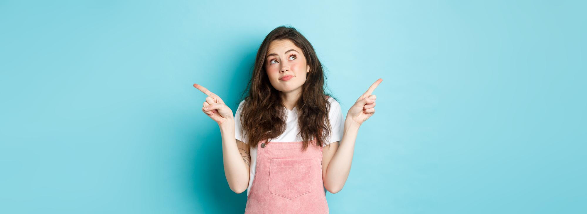 Young woman in pink overalls and white shirt pointing both hands sideways against a blue background.