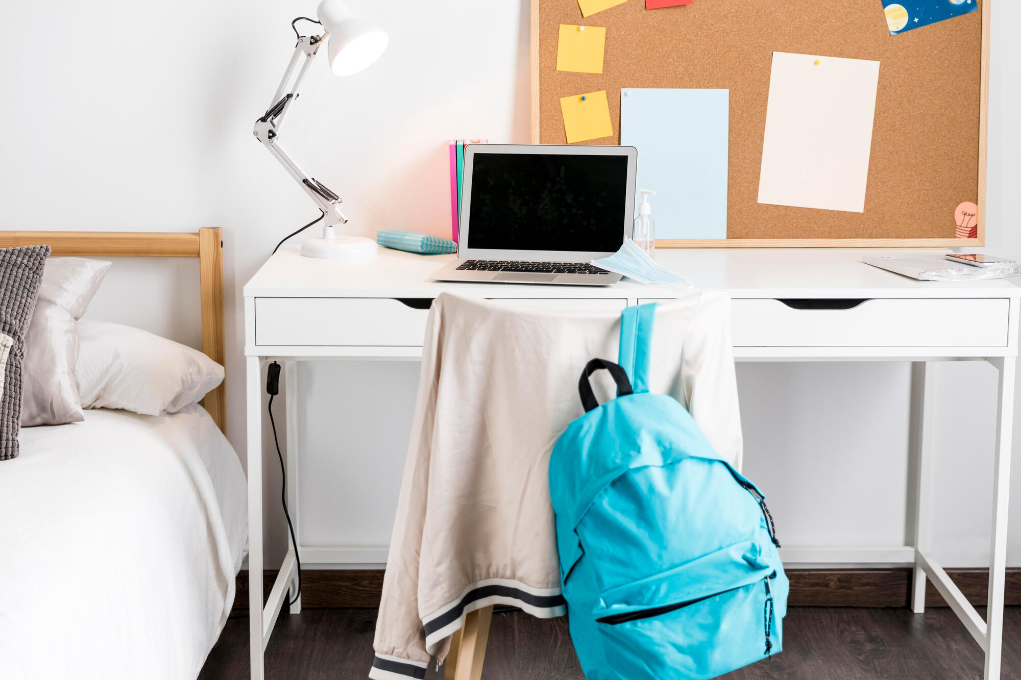 A desk featuring a laptop, a backpack, and a corkboard with notes and reminders pinned on it. 