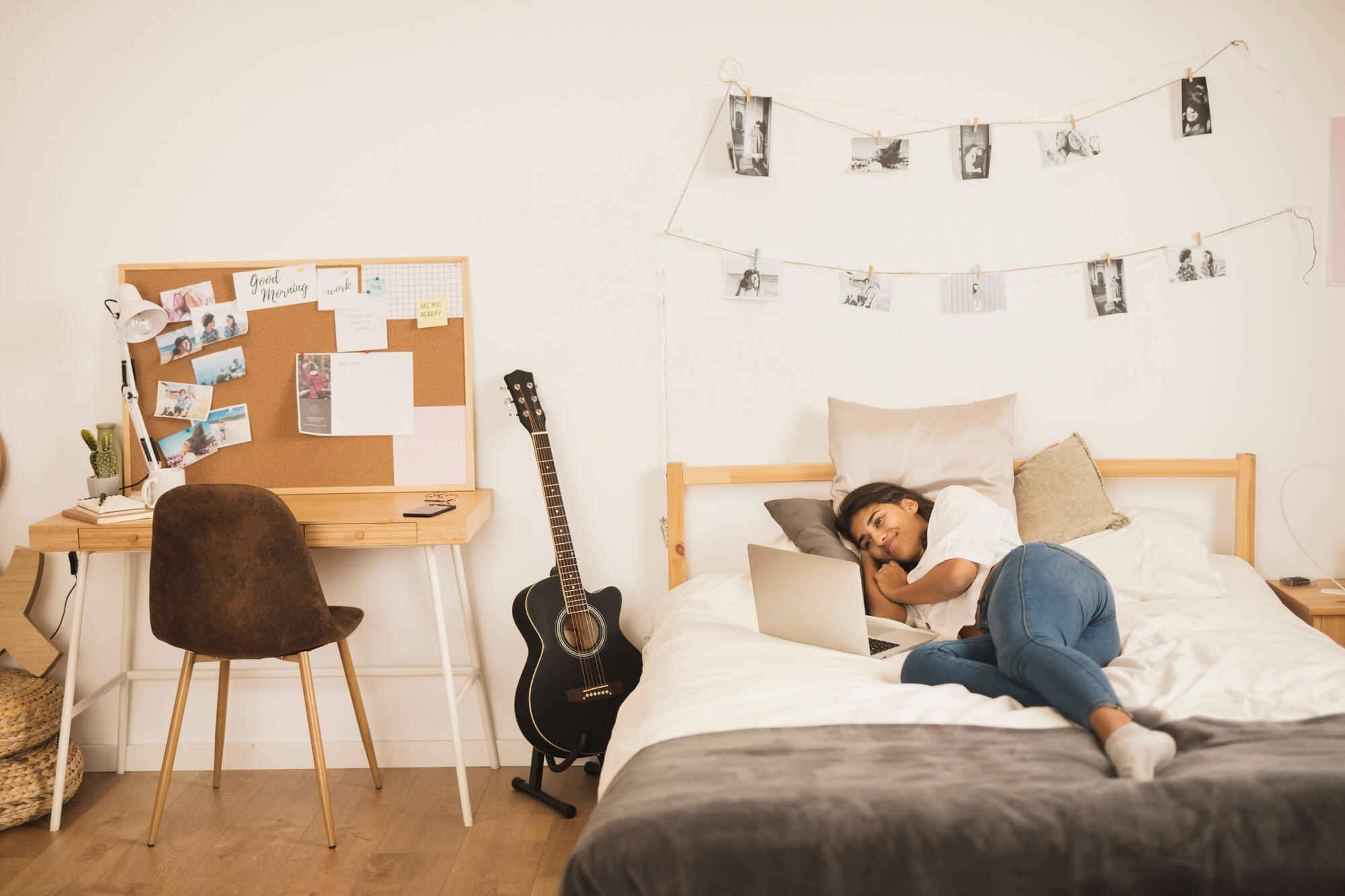 Person lying on a bed using a laptop, with a guitar beside a wooden desk and photos hanging on the wall in a cozy room.
