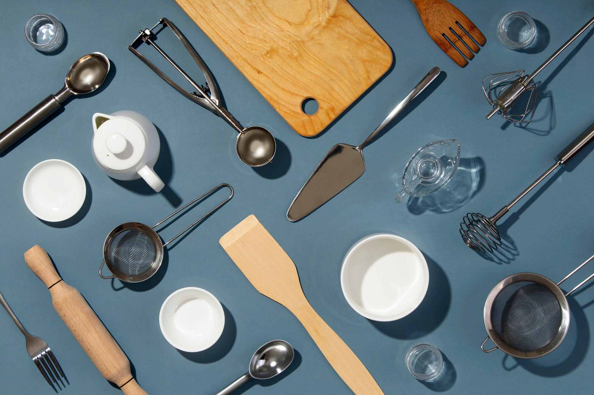 Flat lay of assorted kitchen utensils and tools on a blue surface, including strainers, spoons, a rolling pin, and mixing bowls.