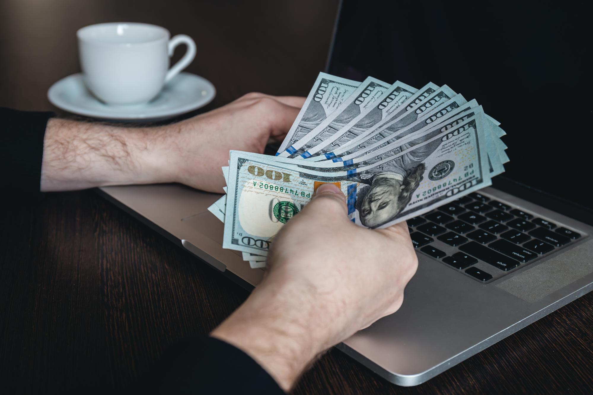 Hands holding multiple US hundred-dollar bills over a laptop keyboard with a white coffee cup in the background on a dark wooden table.