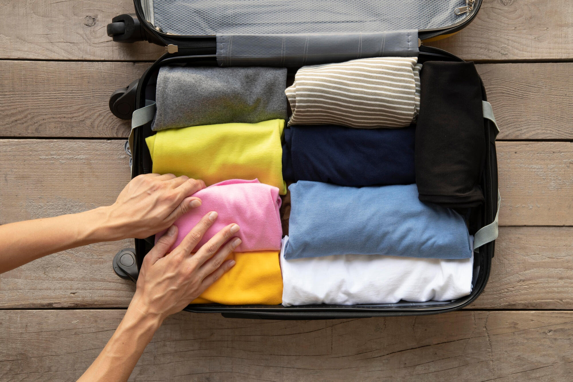 Hands neatly packing a suitcase with folded colorful clothes on a wooden floor background.