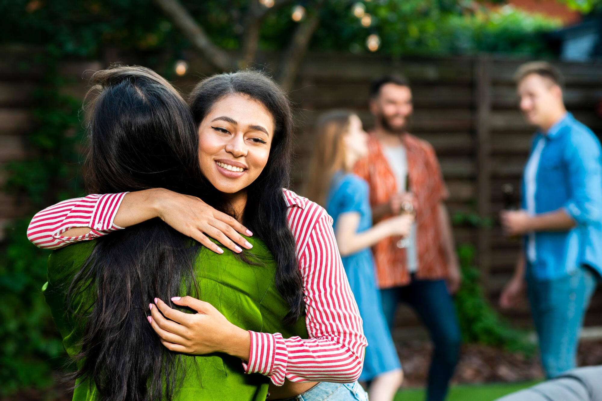 Two women hugging outdoors at a garden party with three people chatting in the background near a wooden fence.