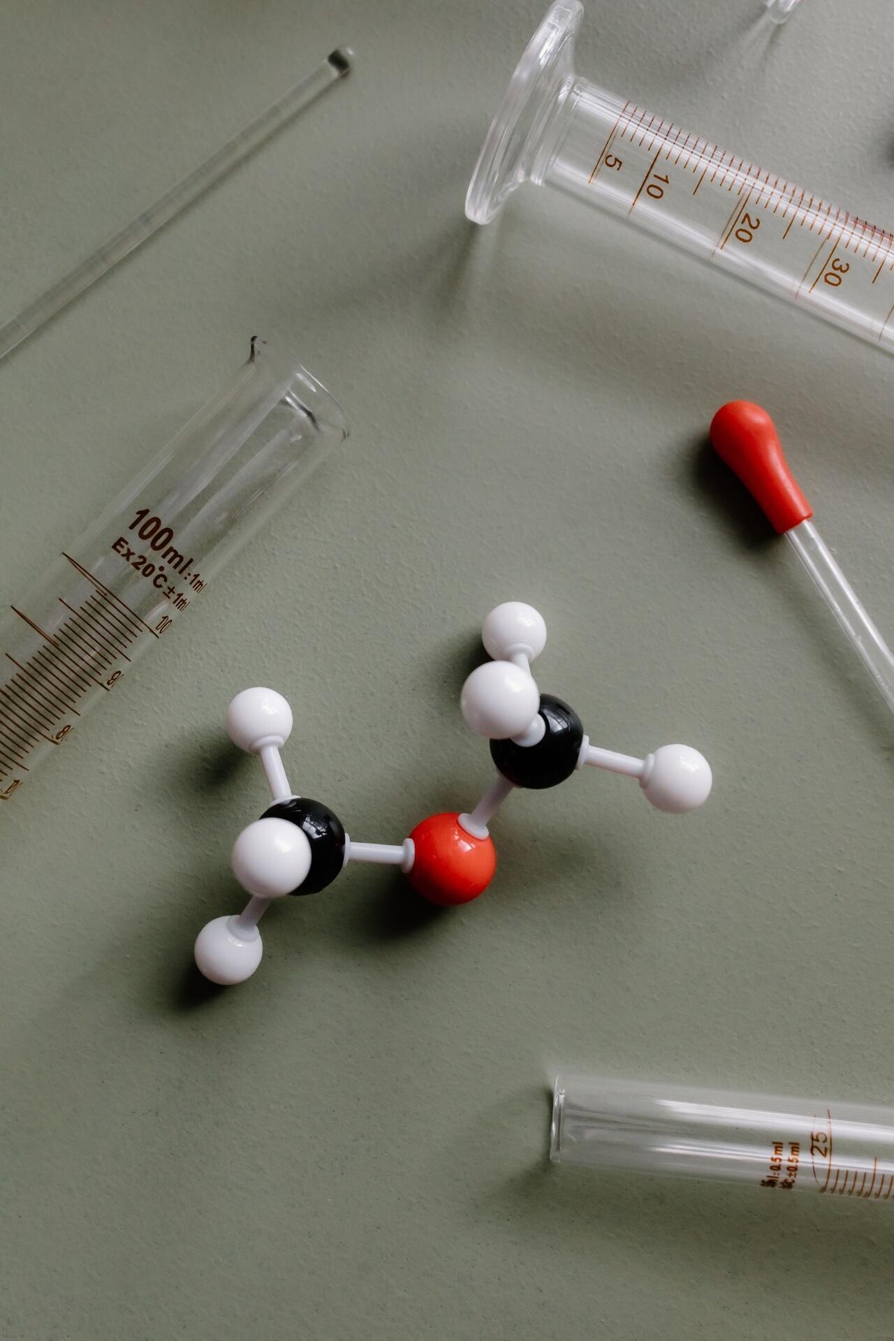 Molecular model surrounded by laboratory measuring equipment, including test tubes, a pipette, and a graduated cylinder.