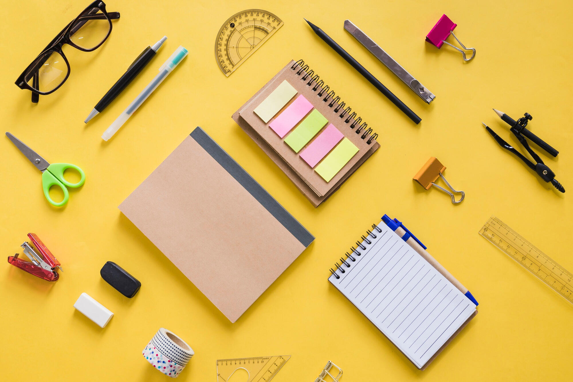 Various stationery items including notebooks, sticky notes, pens, scissors, a protractor, and glasses arranged on a yellow background.