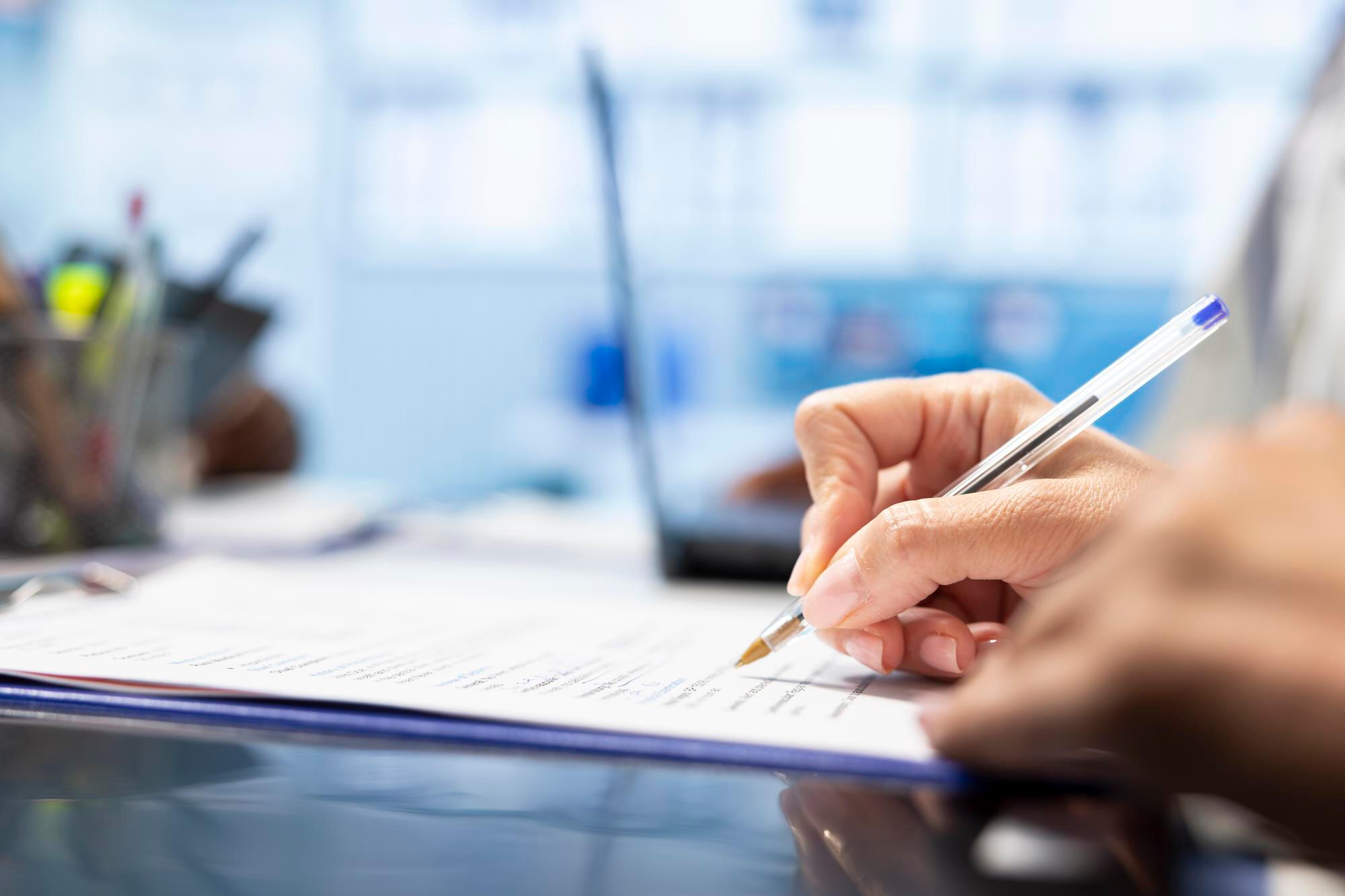 Person's hand holding a pen, filling out or signing a document on a desk with a blurry laptop and office items in the background.