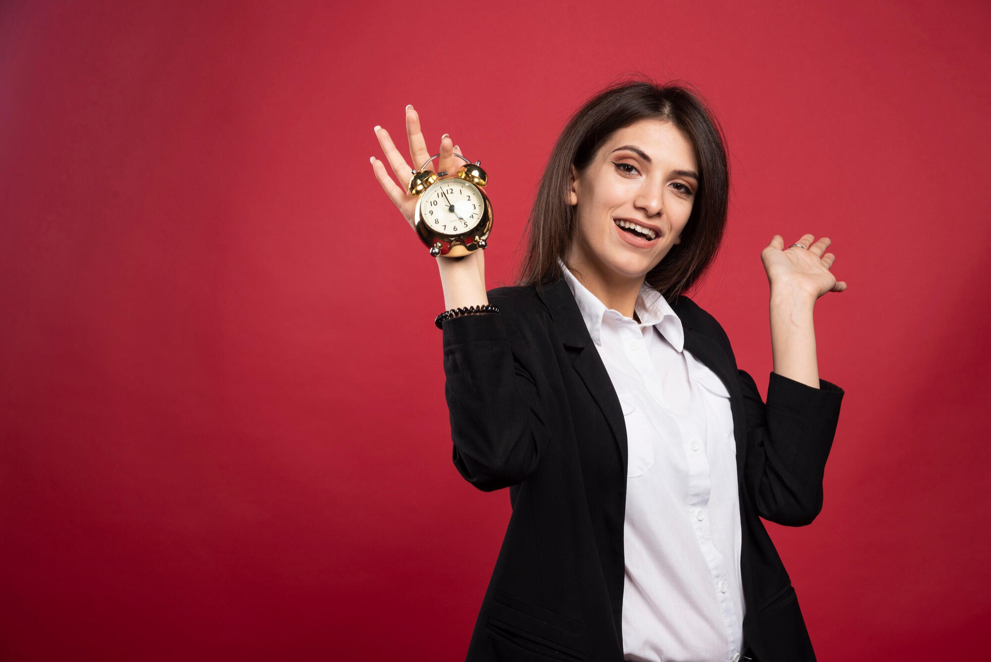 Person in a black blazer holding a small gold alarm clock against a red background, posing with raised hands.