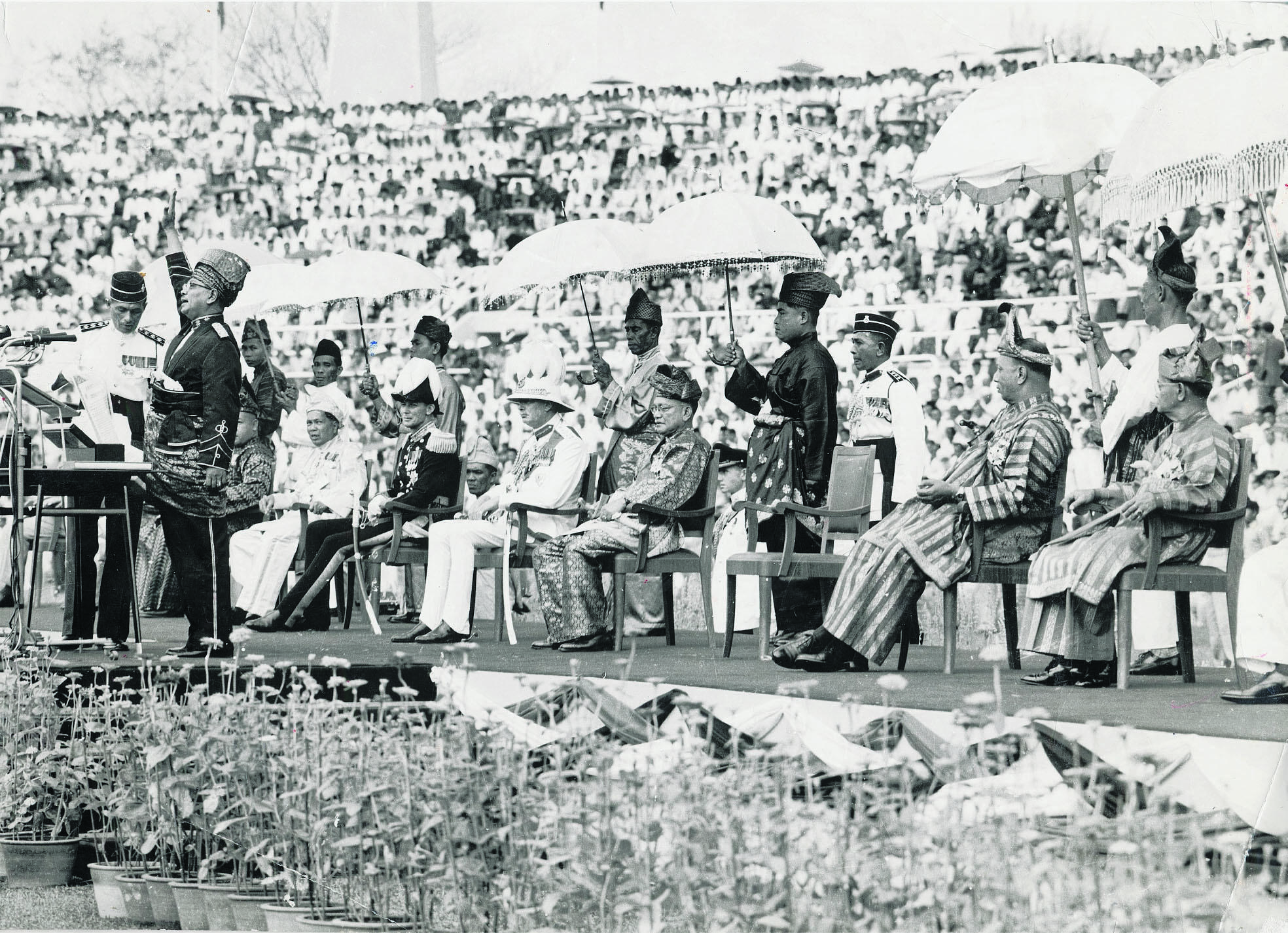Black and white photos depict a large public gathering, with officials seated under umbrellas. The crowd spans a vast area, evoking a formal, historical event.
