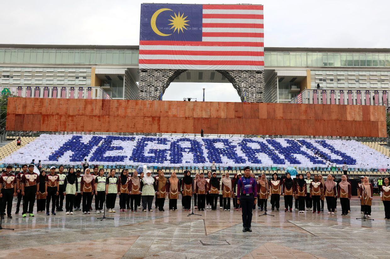 A group of people stand in front of a large Malaysian flag and a display reading "NEGARAKU" in blue, conveying national pride and unity.