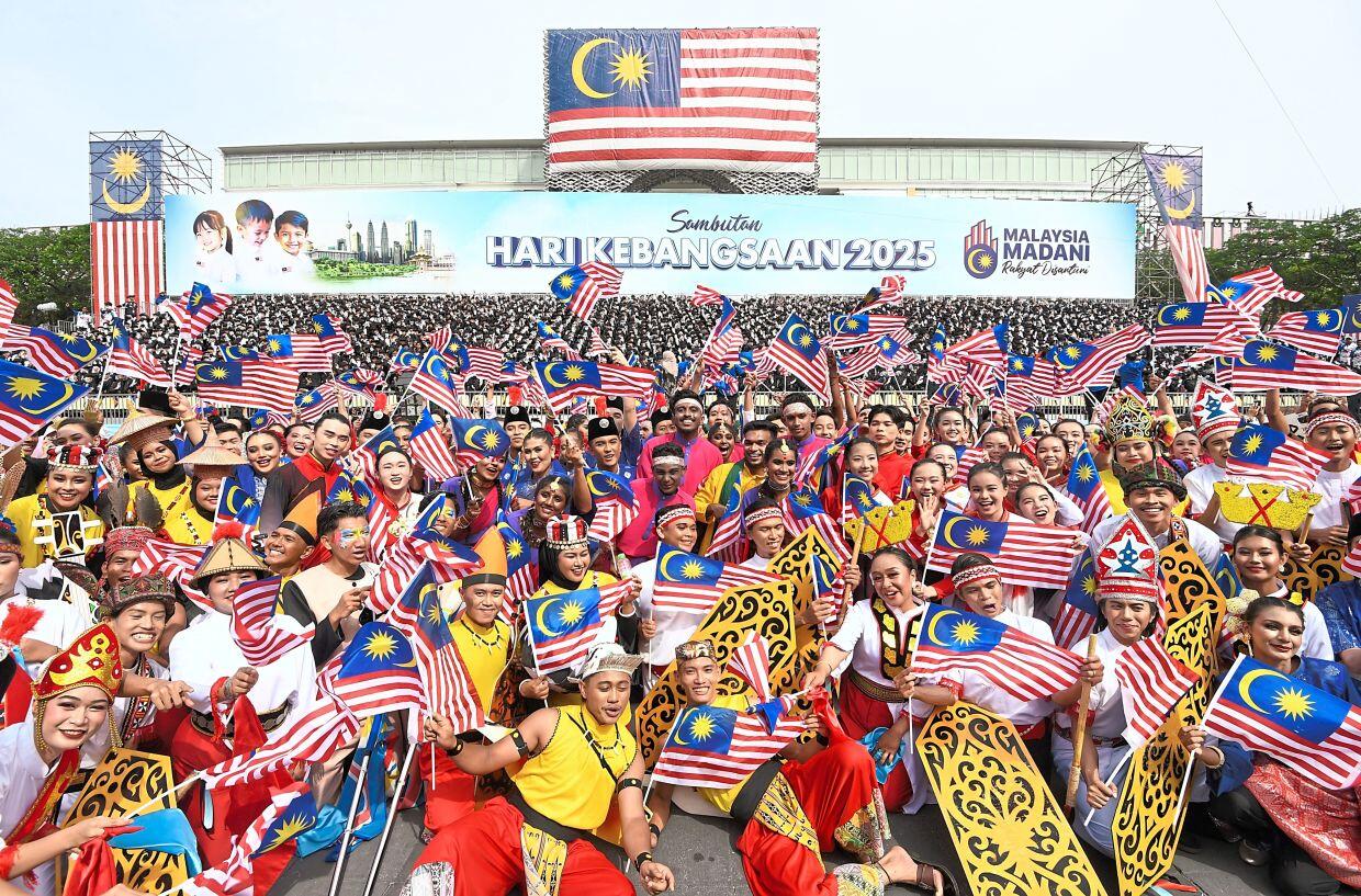 A vibrant crowd dressed in traditional attire celebrates Malaysia's Independence Day 2025, waving Malaysian flags joyfully in front of a large banner.