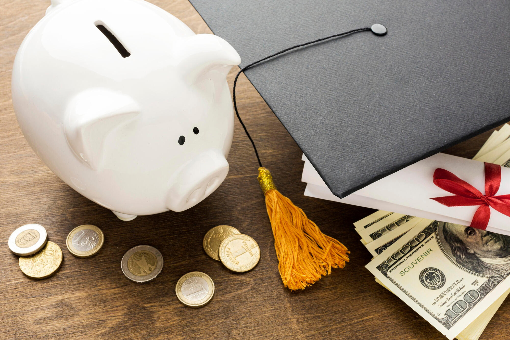 White piggy bank, scattered coins, graduation cap with golden tassel, diploma tied with red ribbon, and stack of hundred-dollar bills on wood table.