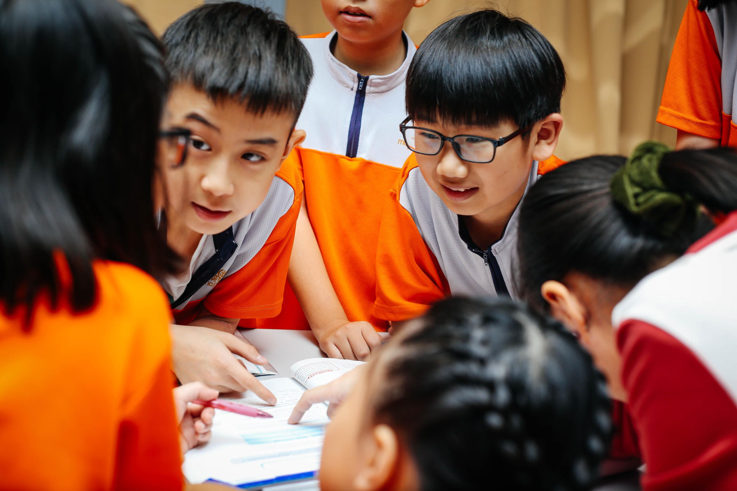 A group of elementary school students in orange and white uniforms gather around a table, looking down at a paper and discussing their schoolwork.