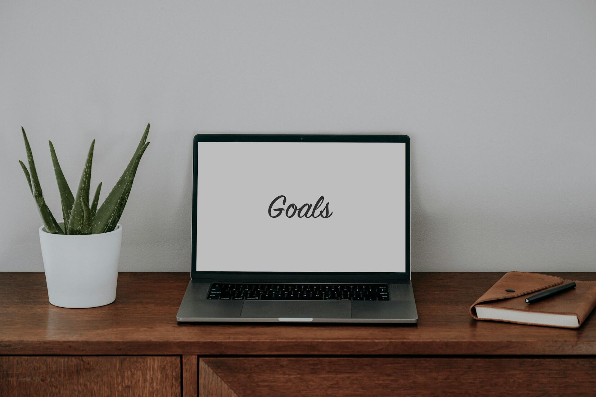 A laptop with the word Goals displayed on its screen sits on a wooden desk next to a potted aloe vera plant and a closed brown leather notebook.