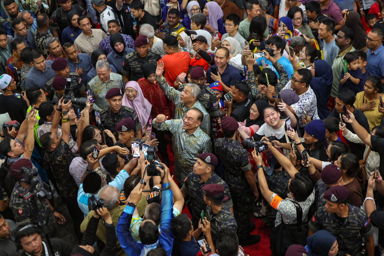 A large crowd of people, including military personnel, gathered around Malaysia's Prime Minister, Dato' Seri Anwar Ibrahim, taking photos and interacting.