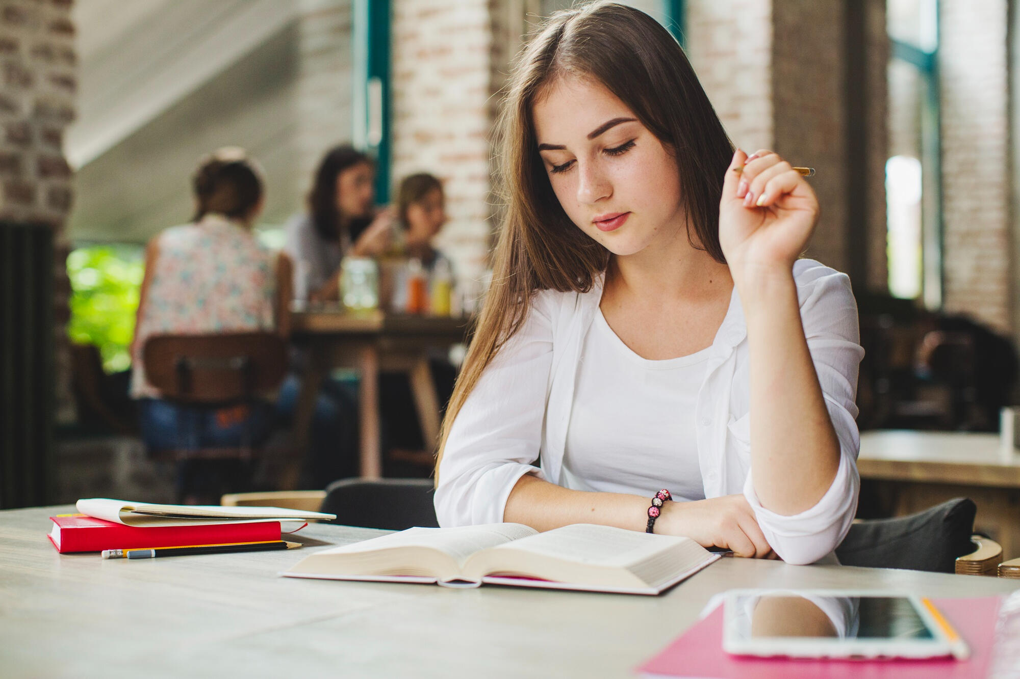 A young woman sits at a table, focused on a book while holding a pen, engaged in writing or studying.