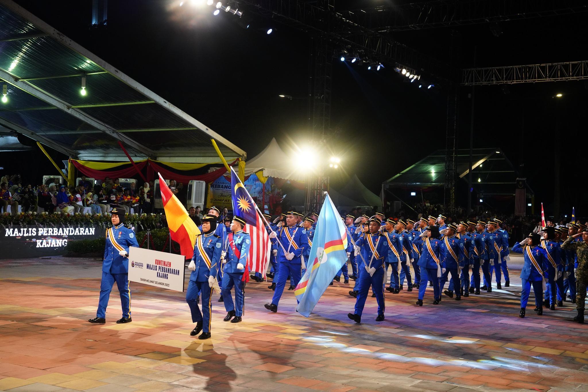 A parade of uniformed participants in blue uniforms and sashes marches at night with flags under bright lights, creating a festive and disciplined atmosphere.