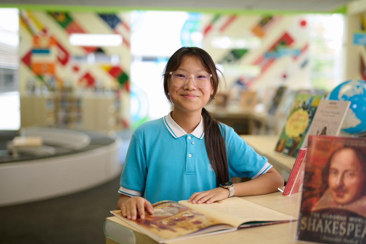 A girl with long hair, wearing glasses and a light blue polo shirt, smiles while sitting at a table in a colorful library. She has an open book in front of her, and a shelf with books is visible in the background.
