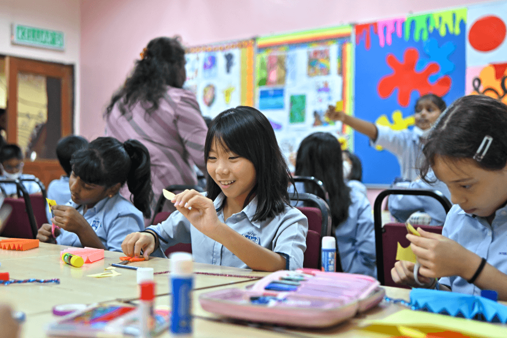 Children in school uniforms sit at a classroom table, smiling and making crafts with paper and glue sticks. Colourful artwork decorates the background walls.