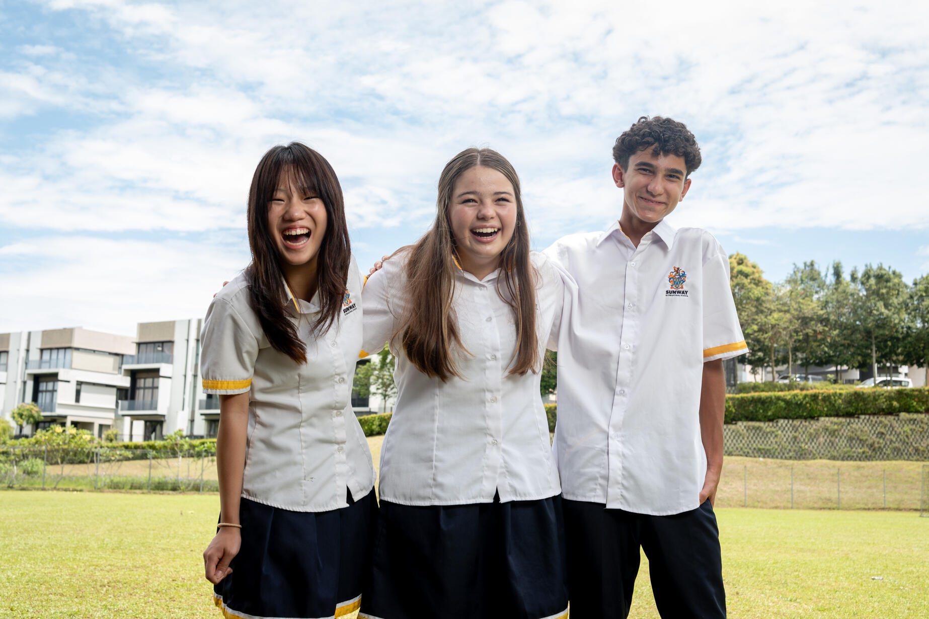 Three students stand close together on a grassy field, smiling. They are wearing matching white school shirts with colored trim and dark skirts or pants. In the background, modern buildings and trees are visible under a partly cloudy sky.