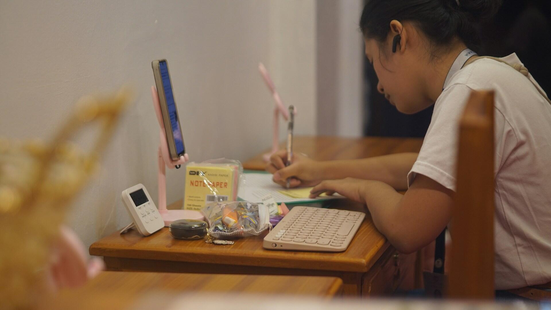 A young woman sits at a wooden desk, writing on a sheet of paper with a pen. A small digital clock and a bag of colorful stationery items are visible on the desk, along with a tablet propped up and a wireless keyboard.