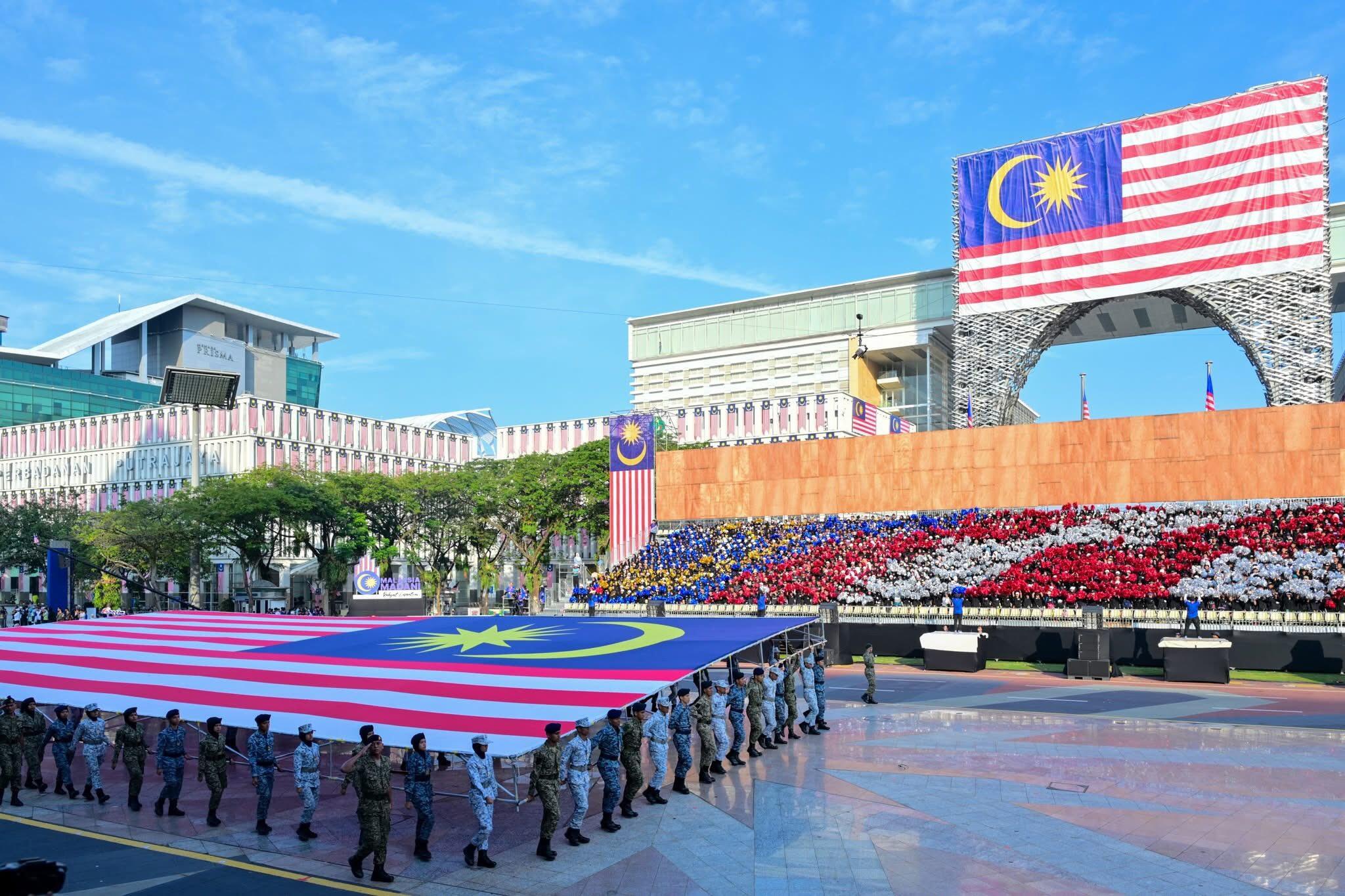 Rows of uniformed individuals carry a large Malaysian flag in front of a modern building. Another flag and a colorful crowd form a patriotic backdrop.