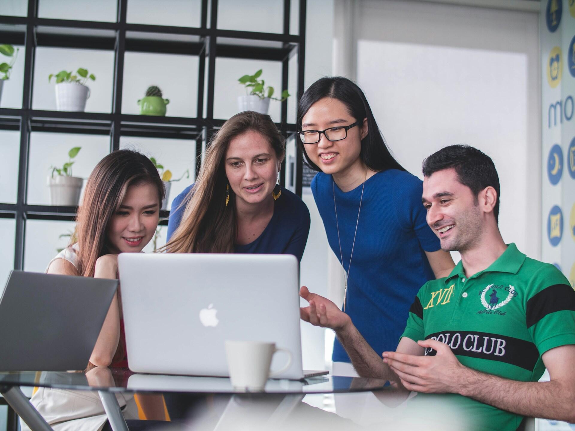 Four people gather around a laptop on a glass table, collaborating on a project. Two women, one with long dark hair and one with long light brown hair, are sitting, while another woman and a man are standing. The background features plants on shelves.