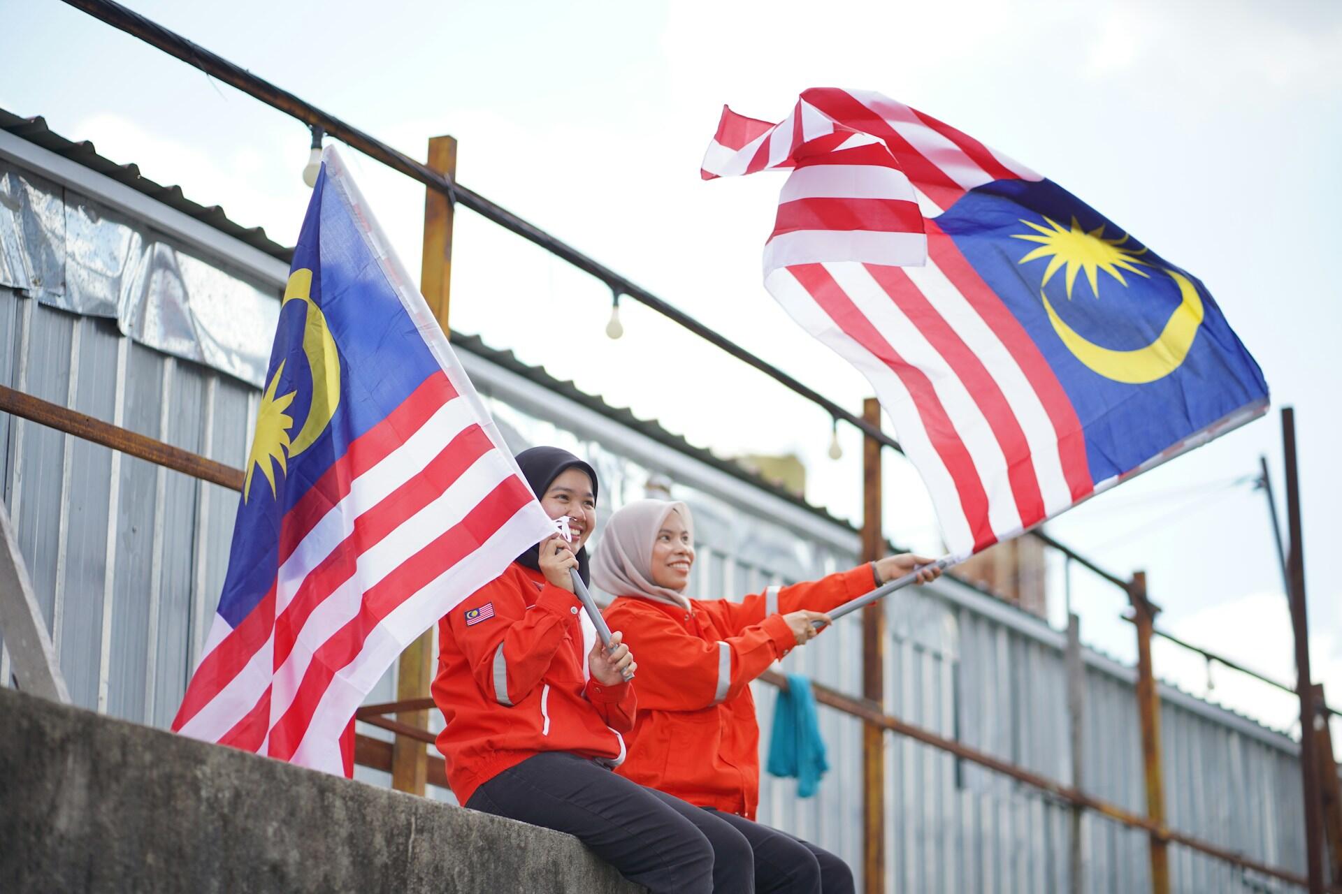 Two people in red jackets wave large Malaysian flags while sitting outdoors on a concrete ledge. They appear joyful, set against a bright, sunny sky.