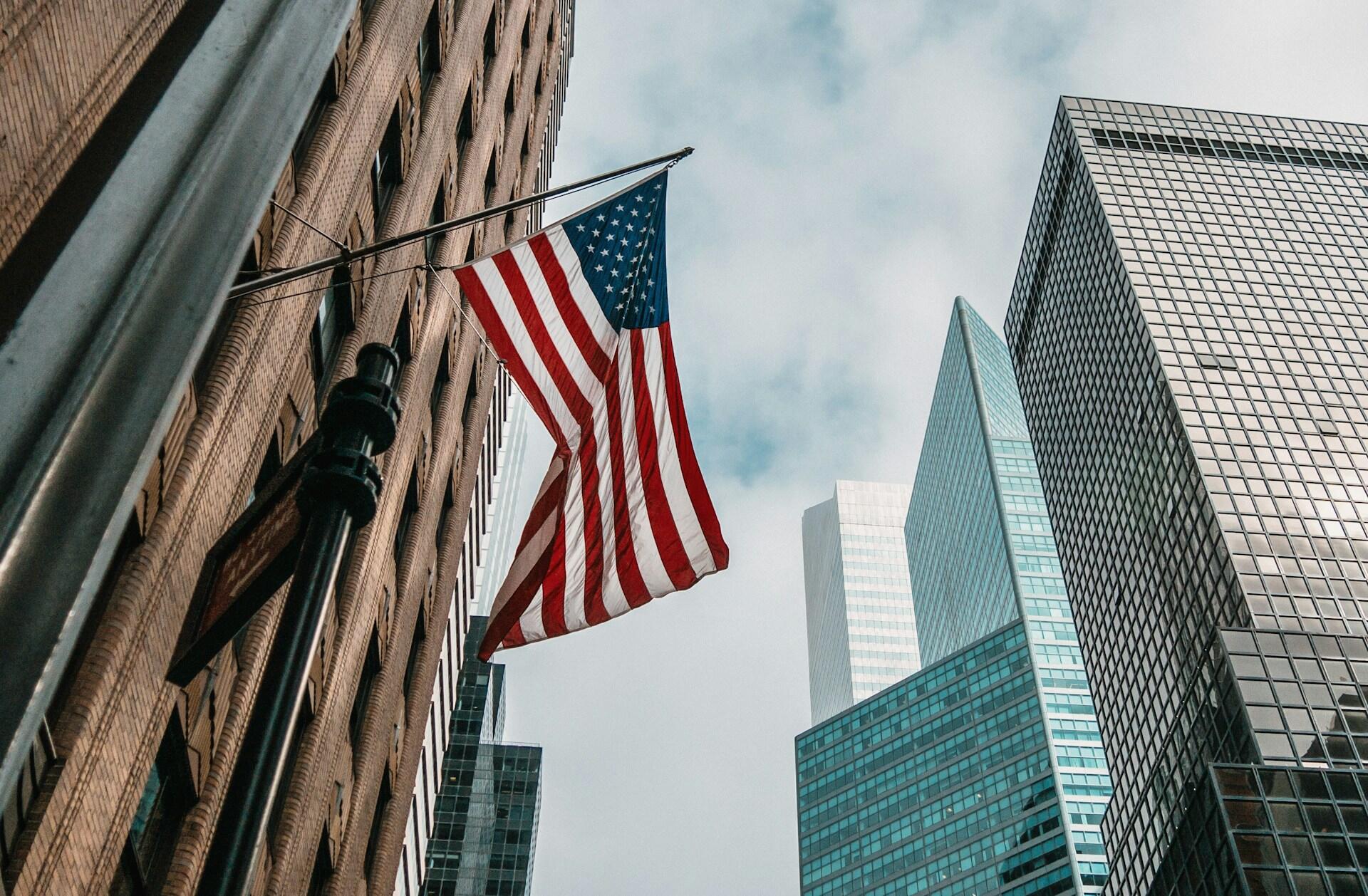 A low-angle view of a United States flag hanging between tall buildings, with a cloudy sky in the background.