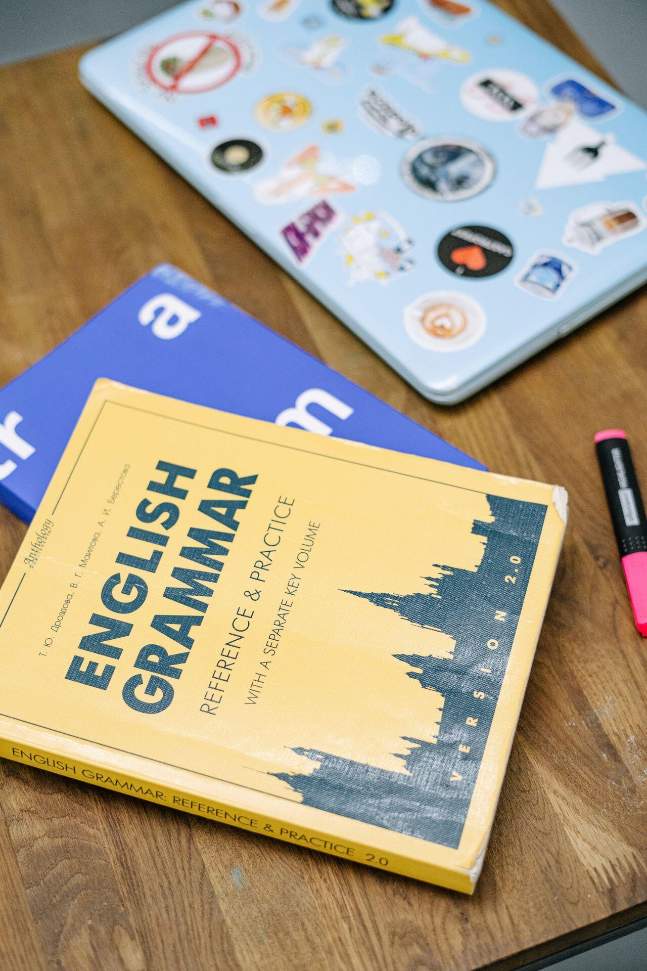 A yellow English Grammar textbook, a blue book, a sticker-covered laptop, and a pink highlighter sit on a wooden table, viewed from above.