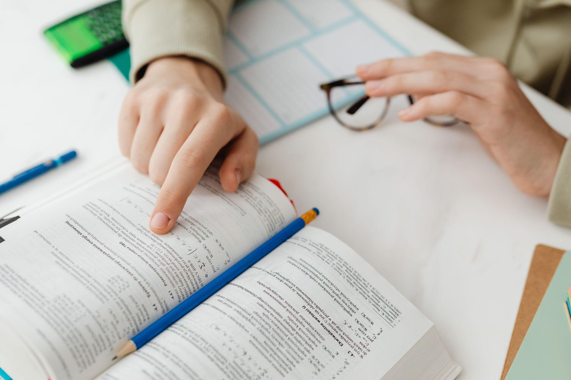 A person studies a technical textbook, pointing at a paragraph with their finger while a blue pencil rests on the open pages at a white desk.