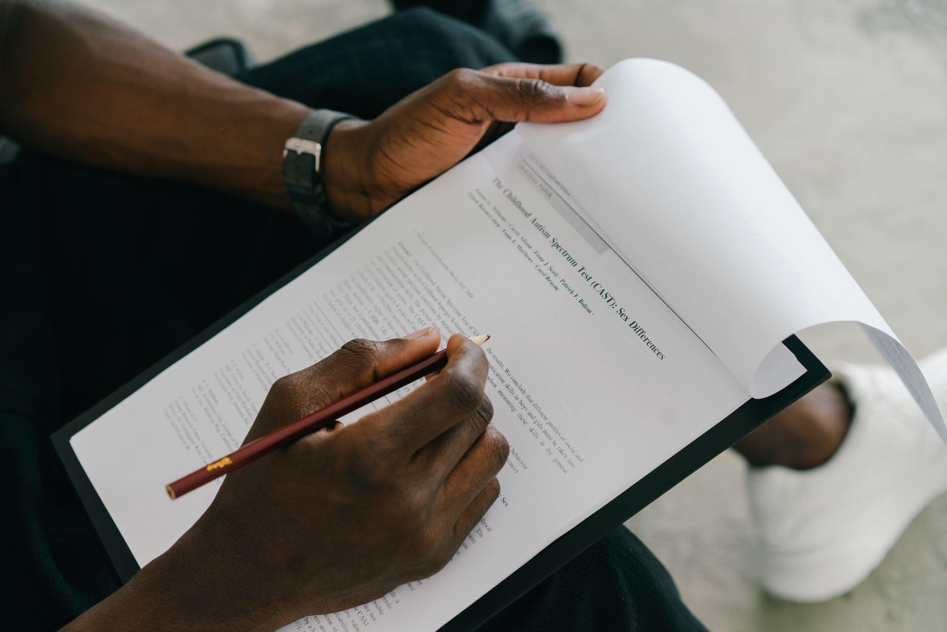 A person with dark skin holds a clipboard with a paper, writing on it with a dark red pencil.