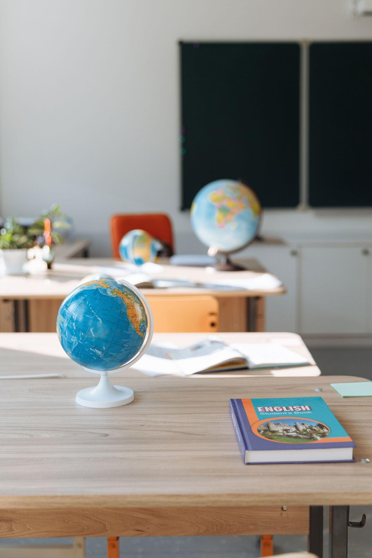 A classroom desk with a blue globe and an English student's book, with more desks, globes, and a chalkboard visible in the blurred background.