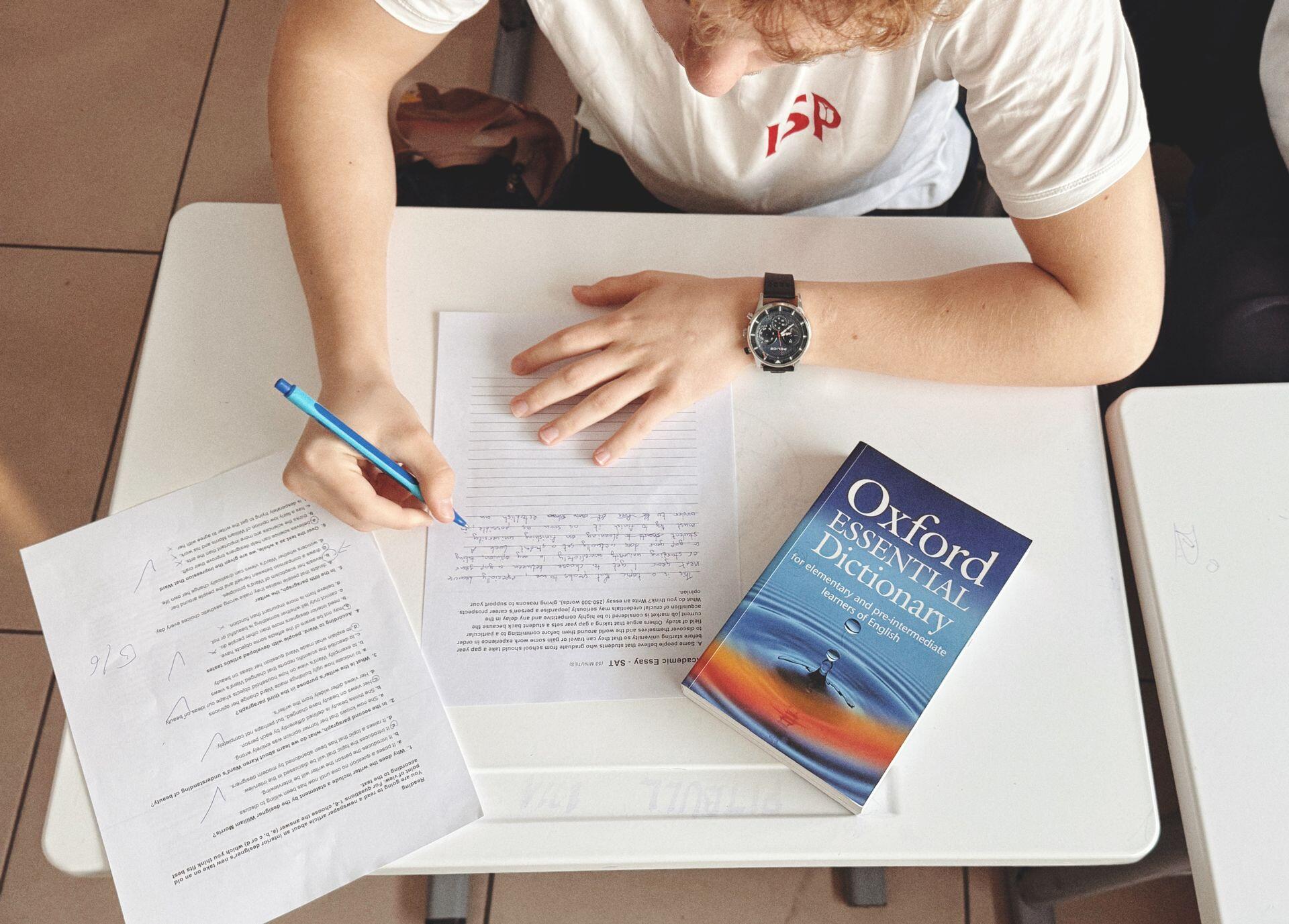 A student in a white t-shirt writes on lined paper at a white desk next to an Oxford Essential Dictionary and a completed multiple-choice test.
