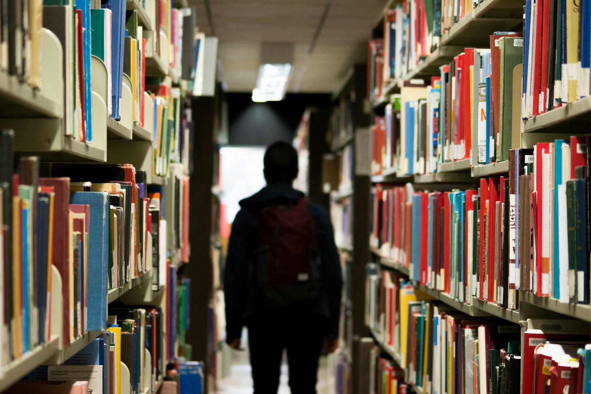 A person with a backpack walks down a narrow aisle between tall, wooden bookshelves filled with many colorful books in a brightly lit library.