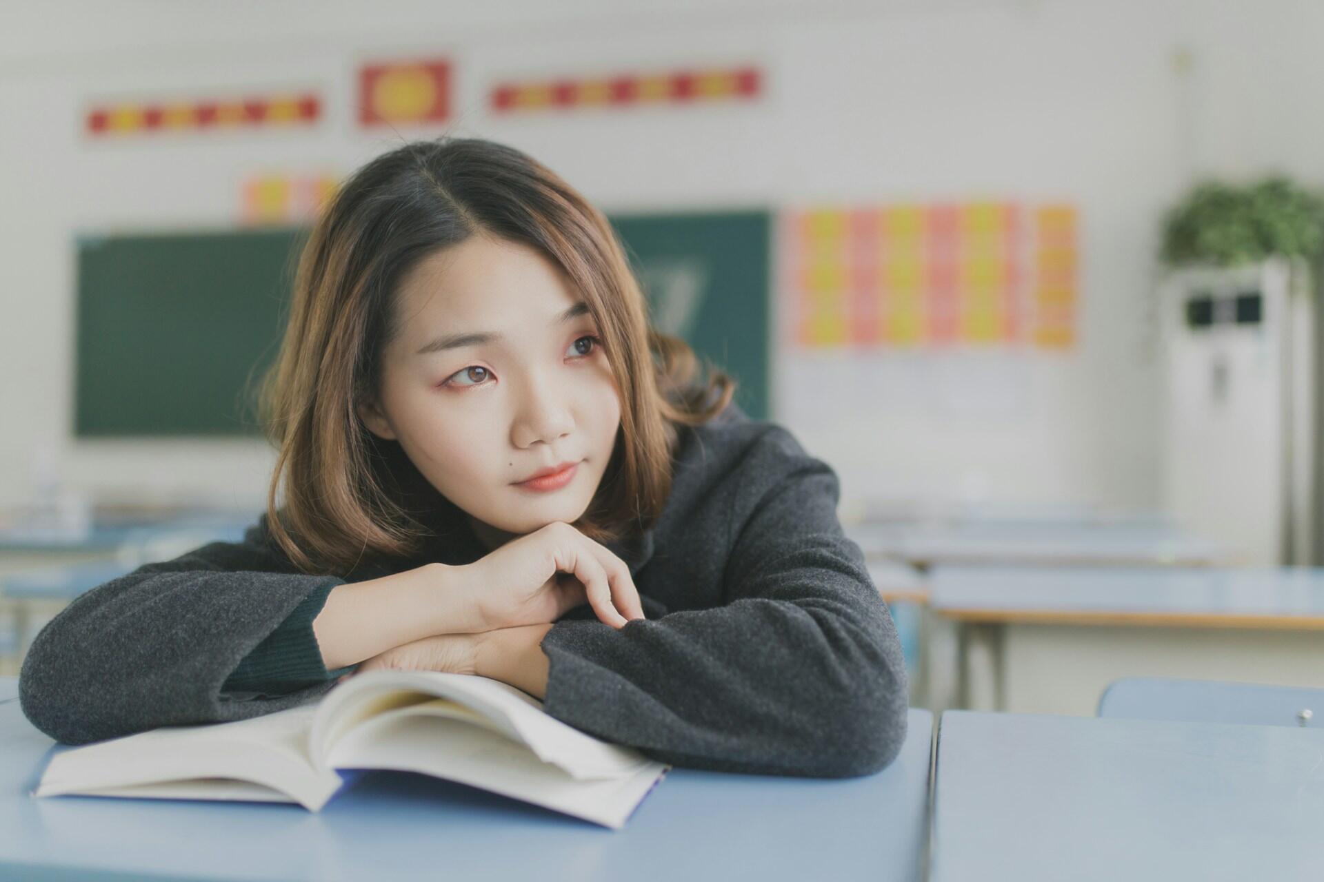 A student in a grey dress sits at a table, resting her hands, pausing to think.