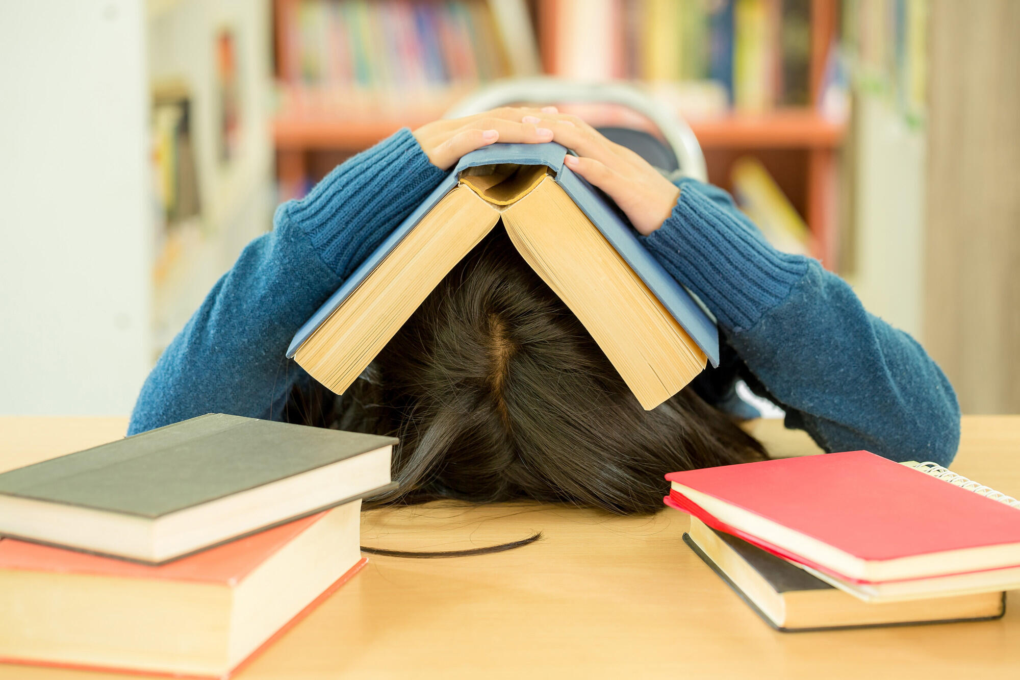 Person with head on desk covered by an open book, surrounded by several closed books, in a library or study setting.