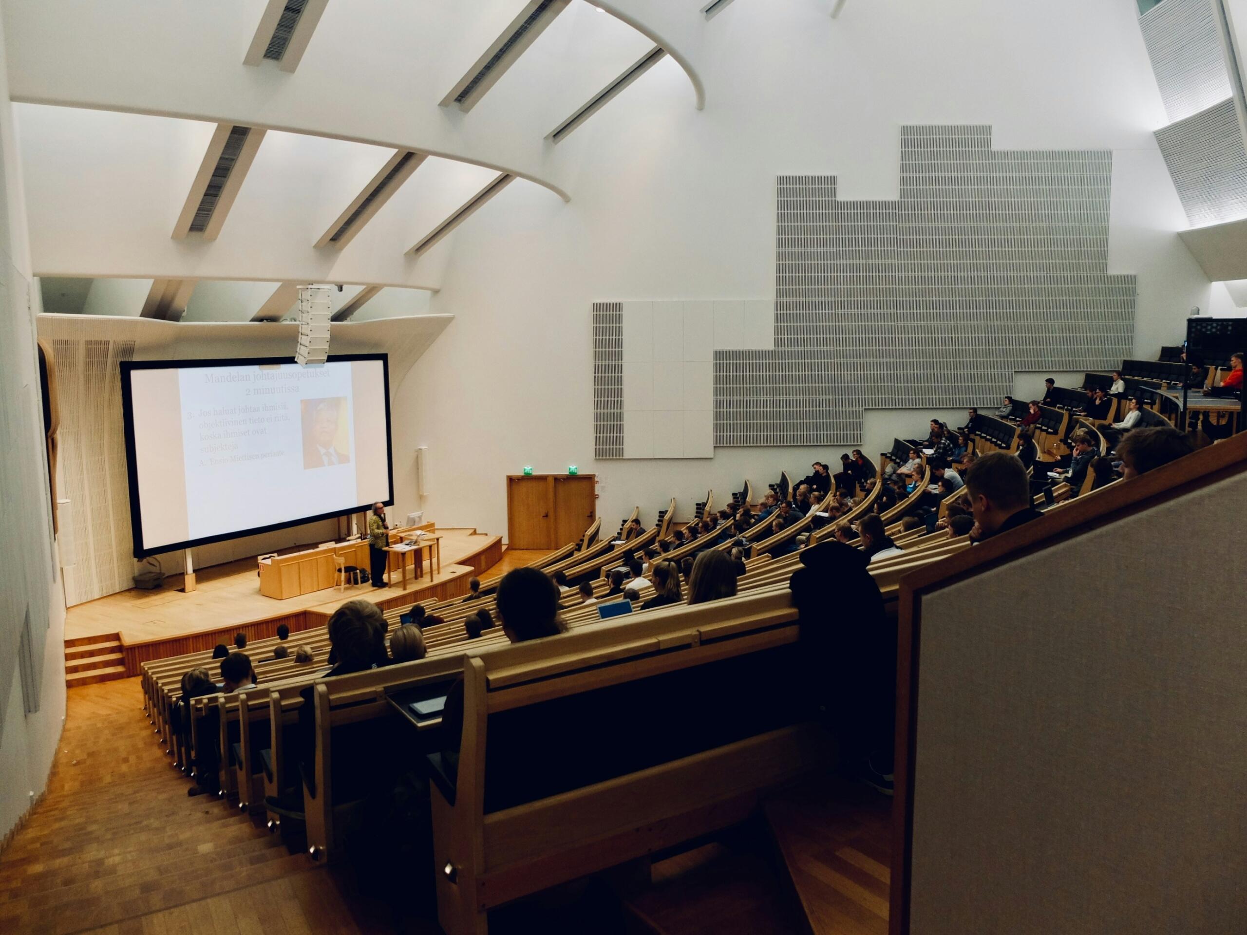 A row of university students seated at desks with laptops in a bright modern lecture hall.