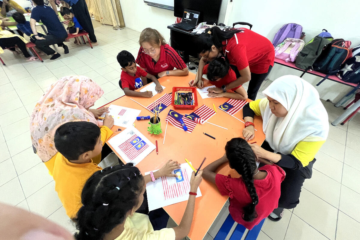 Children and teachers gather around a table, drawing and coloring Malaysian flags with vibrant pencils. The atmosphere is lively and educational.