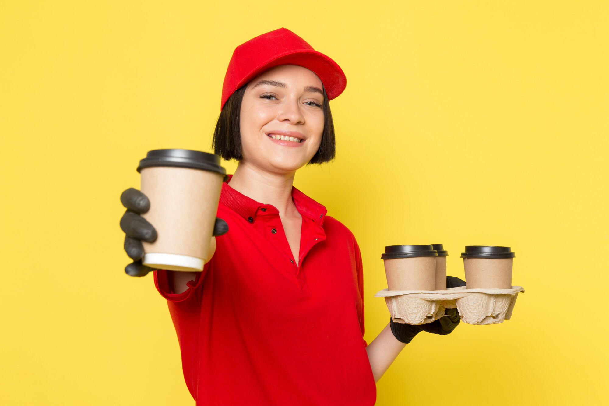 Woman in red uniform and cap holding three takeaway coffee cups, smiling against a yellow background.