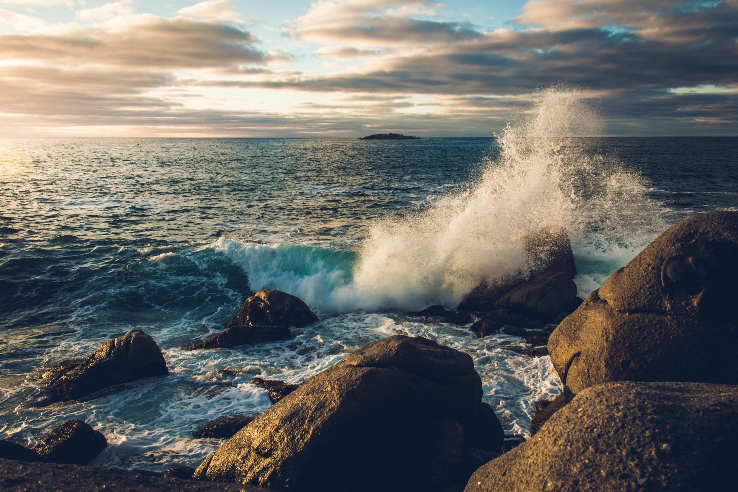 The sun sets behind crashing waves on rocks, highlighting the beauty of the ocean's power.
