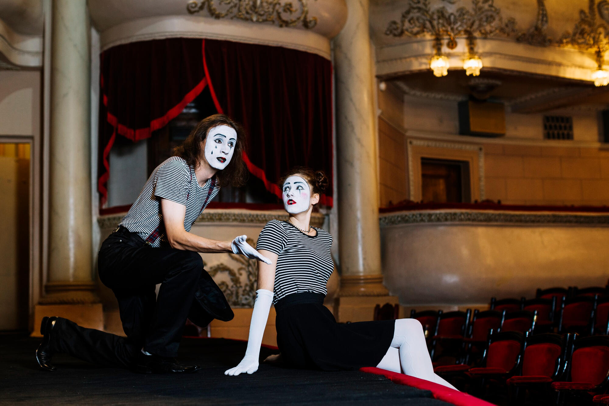 Two mime performers in striped shirts and white face paint pose onstage inside an ornate theater.