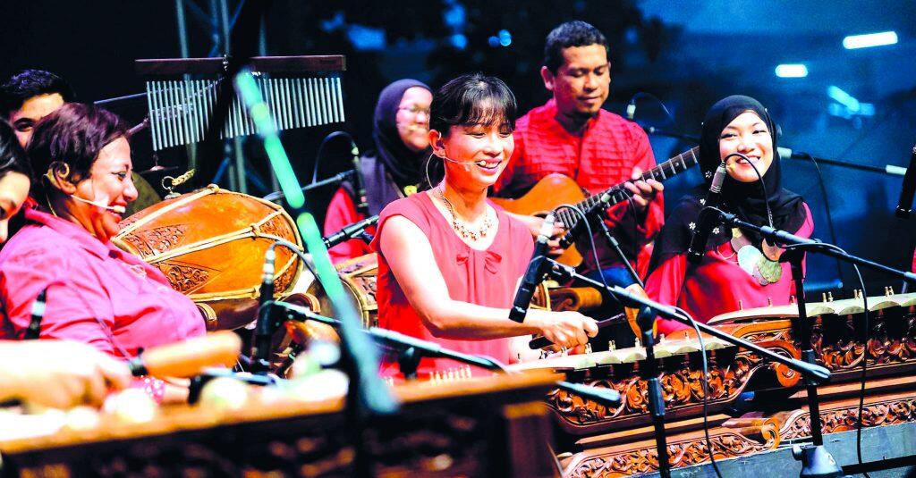 A musical ensemble performs on stage with traditional instruments, including a gamelan, while wearing red clothing under bright stage lighting.