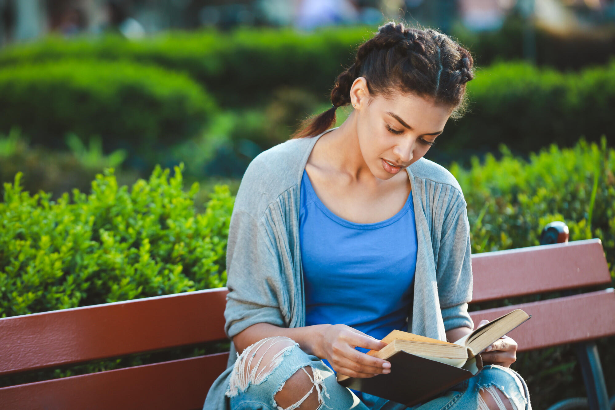 Young woman sitting on a park bench, reading an open book surrounded by greenery.