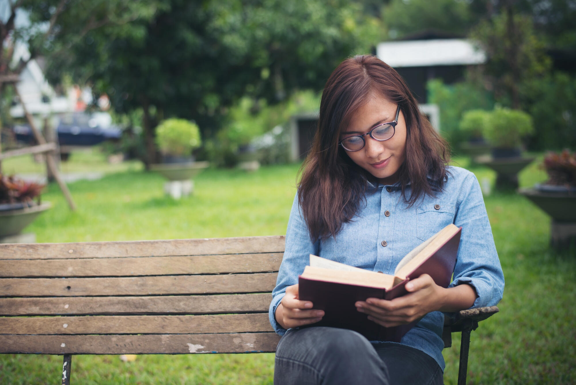 A woman reading a book while seated on a bench, surrounded by a peaceful outdoor environment.