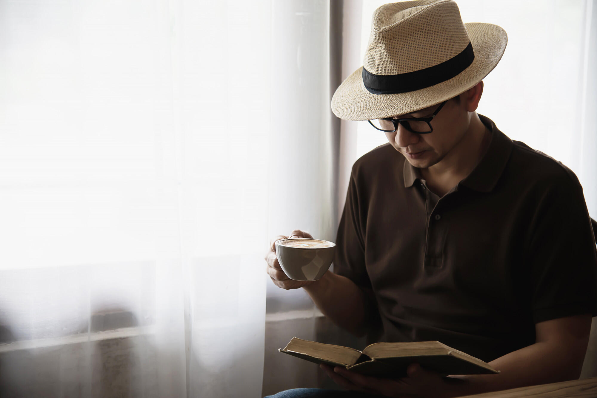 A man with glasses and a hat enjoys a book and coffee at a cozy café setting.