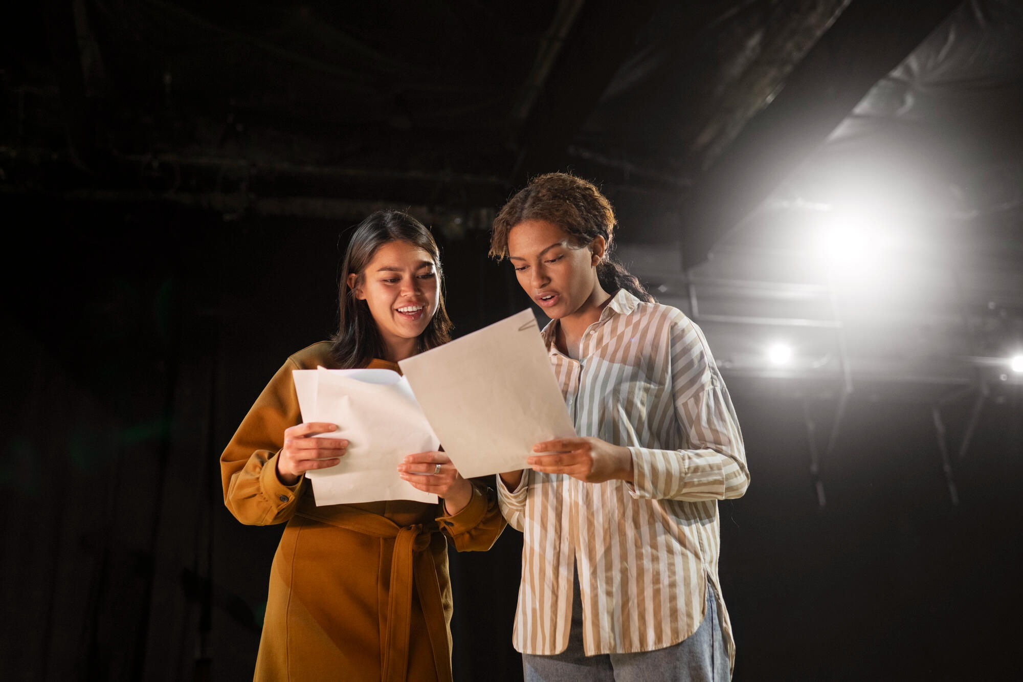 Two women backstage review printed papers together under bright stage lights.