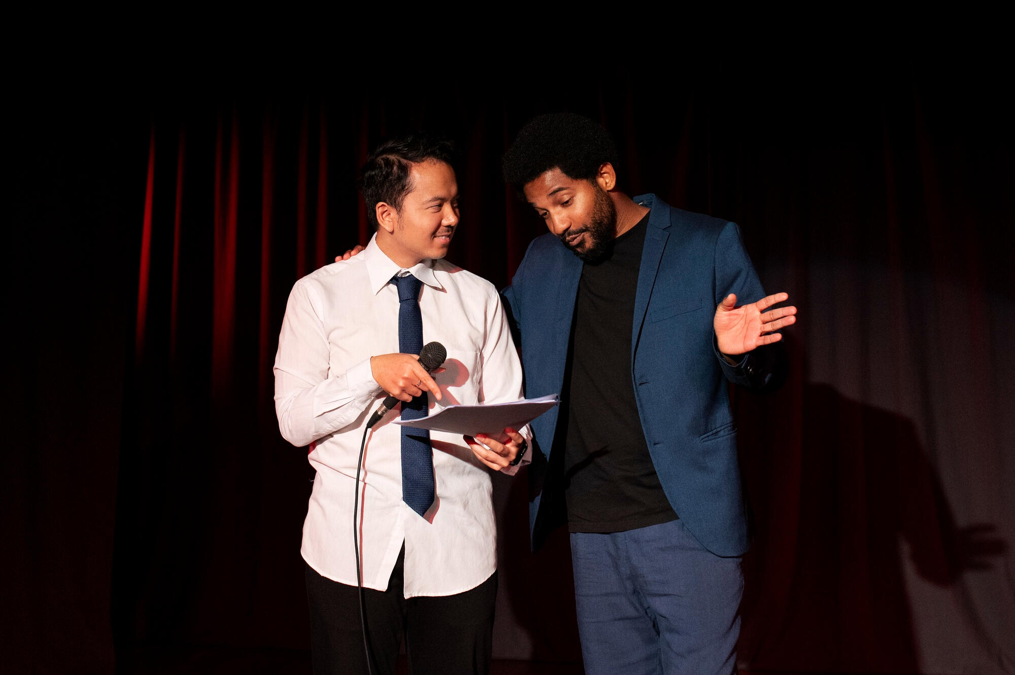 Two men stand onstage before red curtains; one holds a microphone and notes while the other gestures during a presentation.