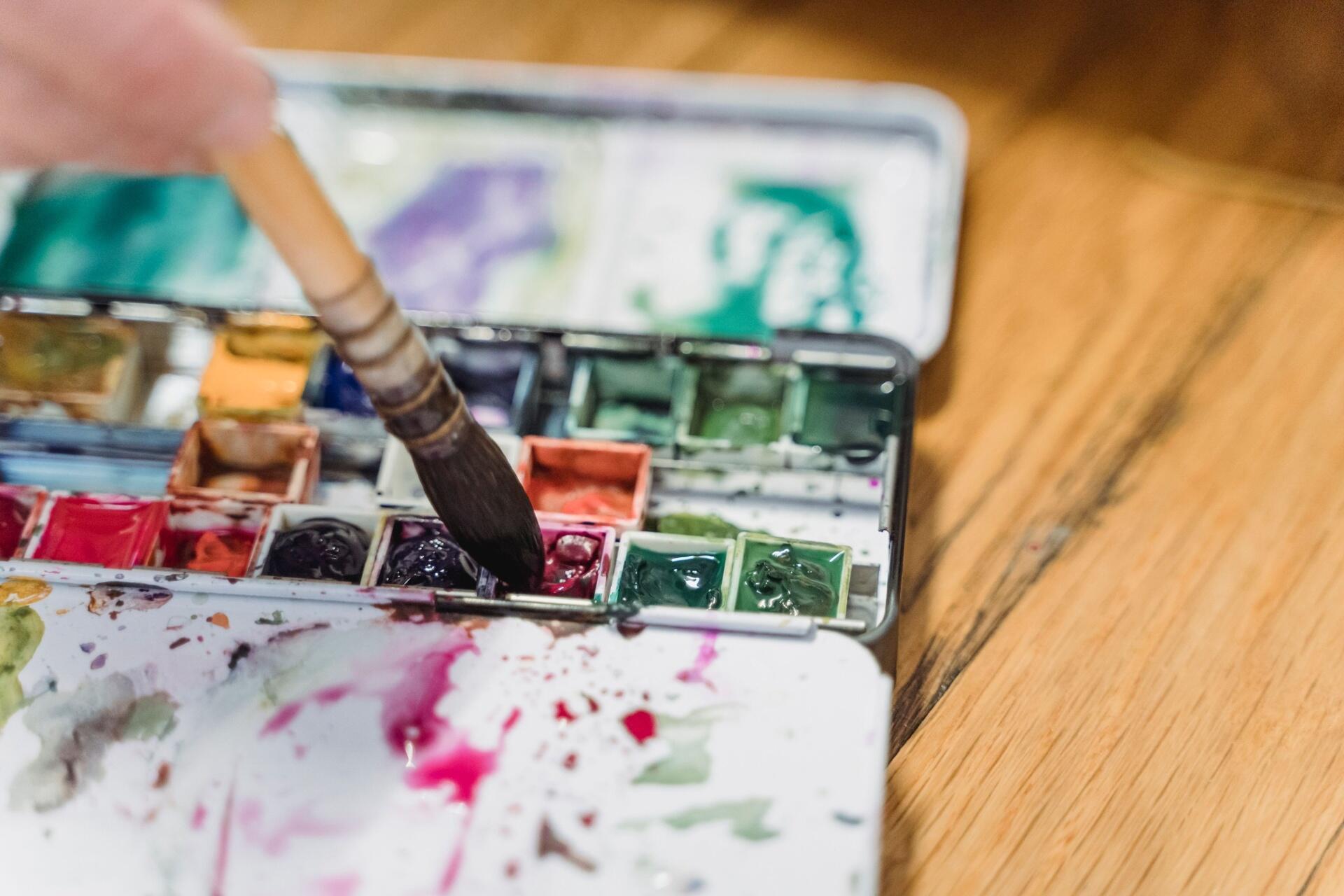 Paintbrush dipped into a watercolour palette containing red, green, blue and other colours on a wooden table.